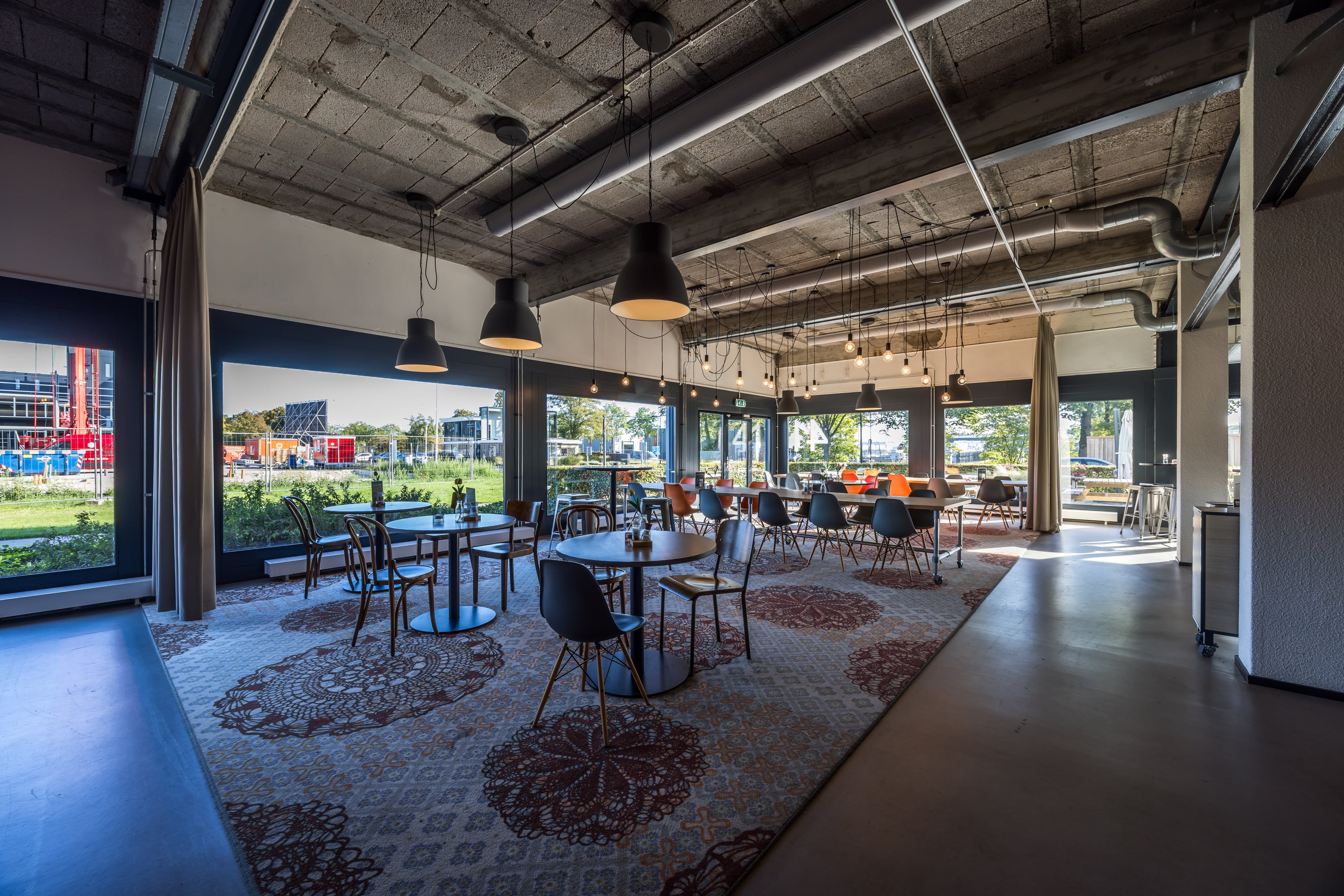 Restaurant met ronde tafels, hanglampen en grote ramen langs de gevel
